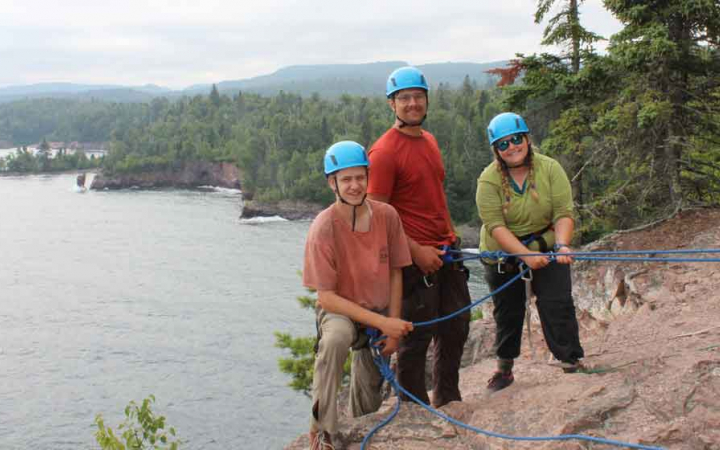Three people wearing safety gear and secured by ropes stand on a rocky cliff and smile at the camera. They are high above a lake. 